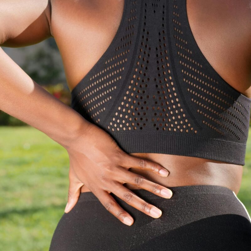 A woman in sportswear gripping her lower back, potentially indicating discomfort, set in an outdoor environment.