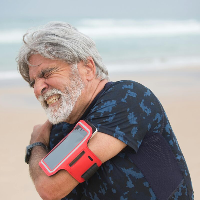Elderly man grimacing from shoulder pain during seaside walk, showcasing health issue outdoors.